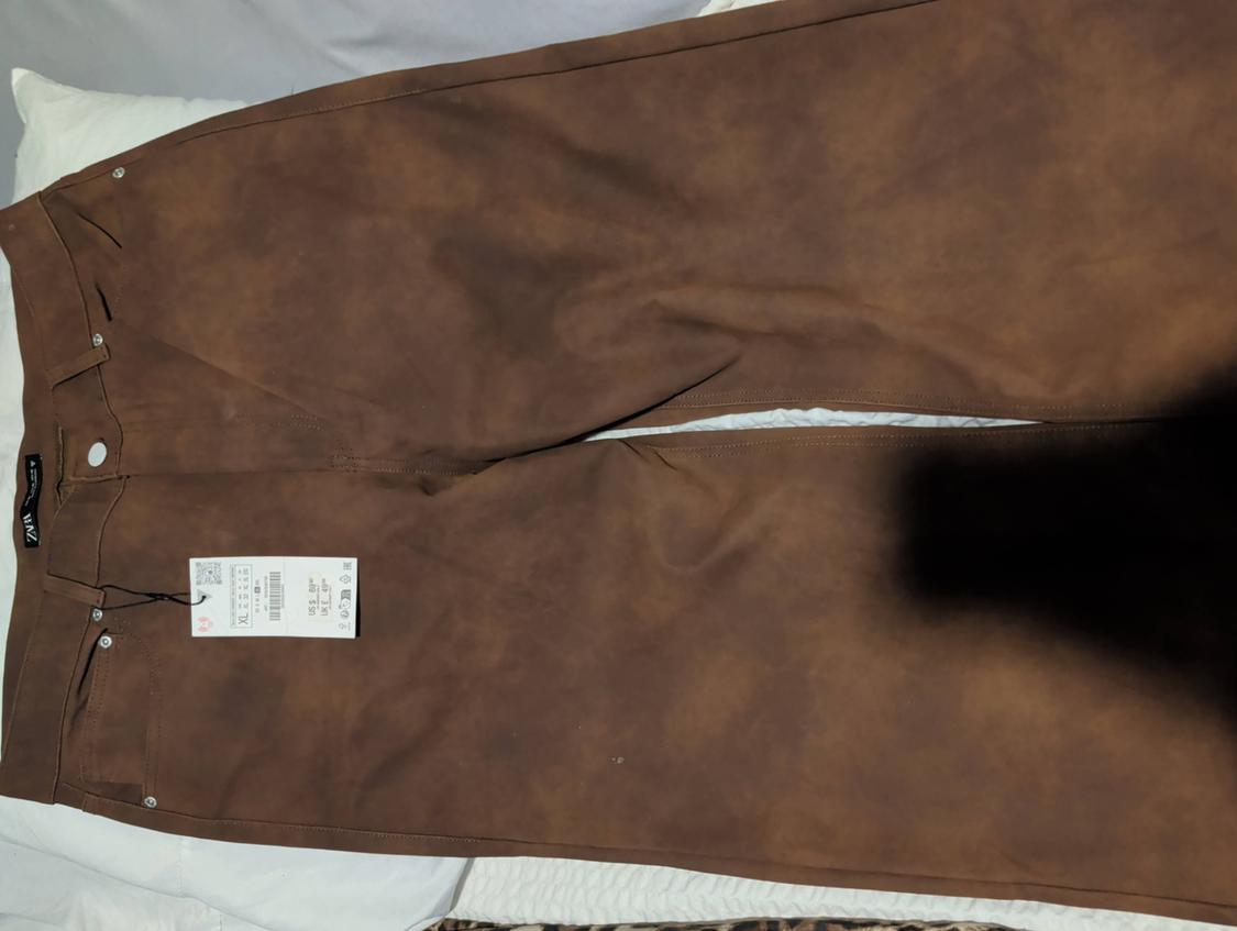 Straight-cut brown pants on a white bed, with a visible hangtag on the left side.