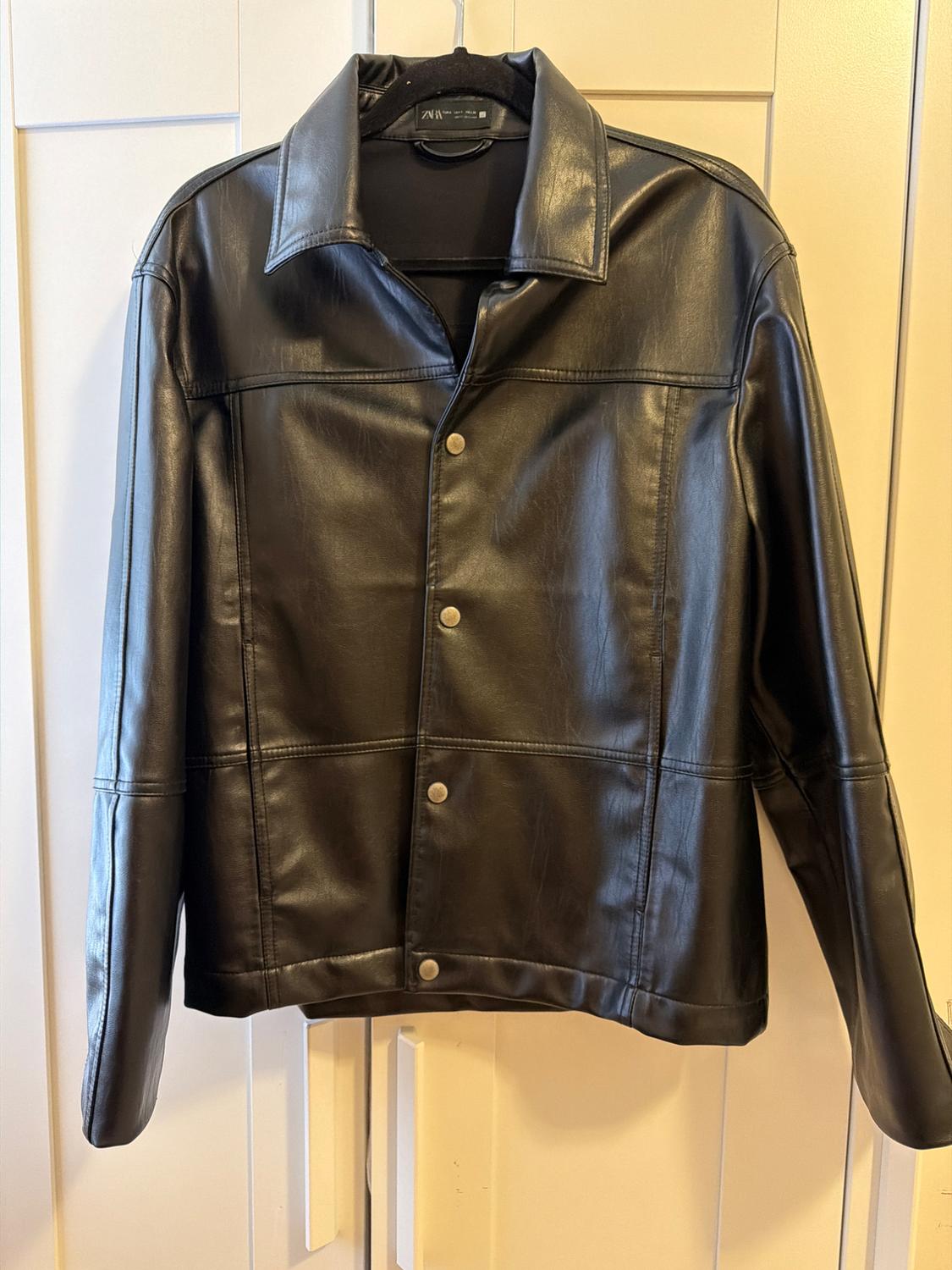 Black jacket with a collar and front buttons, hanging on a black coat hanger against a white door. Interior label with the text ZARA.