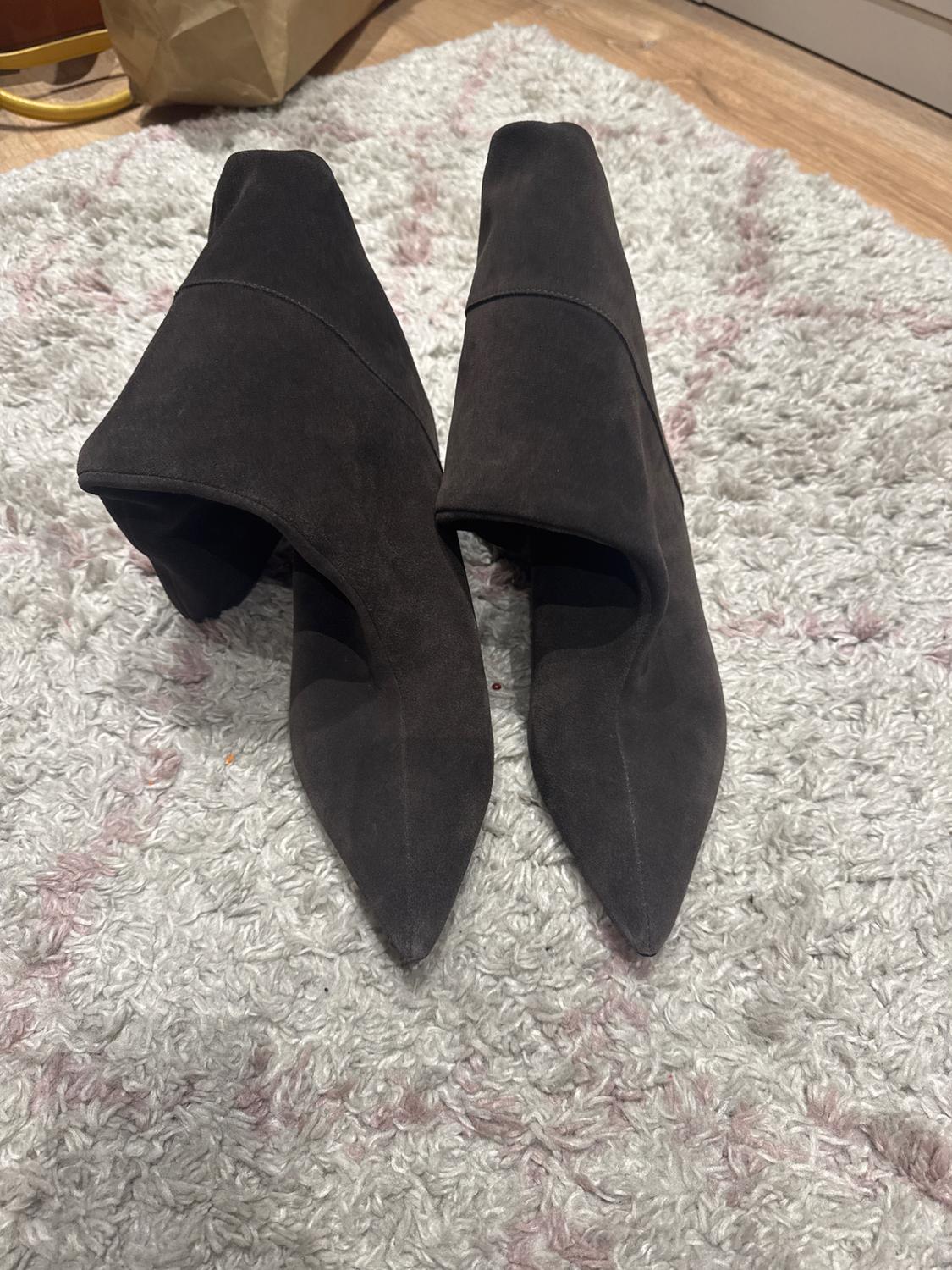 Black high-heeled, pointed-toe boots on a white rug with pink details, on a light wooden floor.
