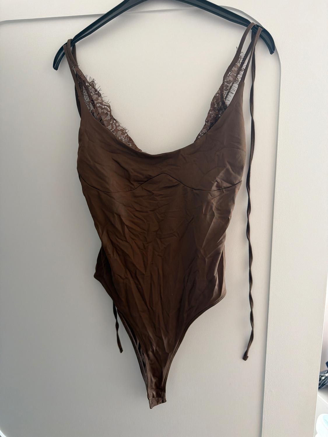 Brown V-neck bodysuit with lace detail and spaghetti straps, hanging on a black hanger against a white background.