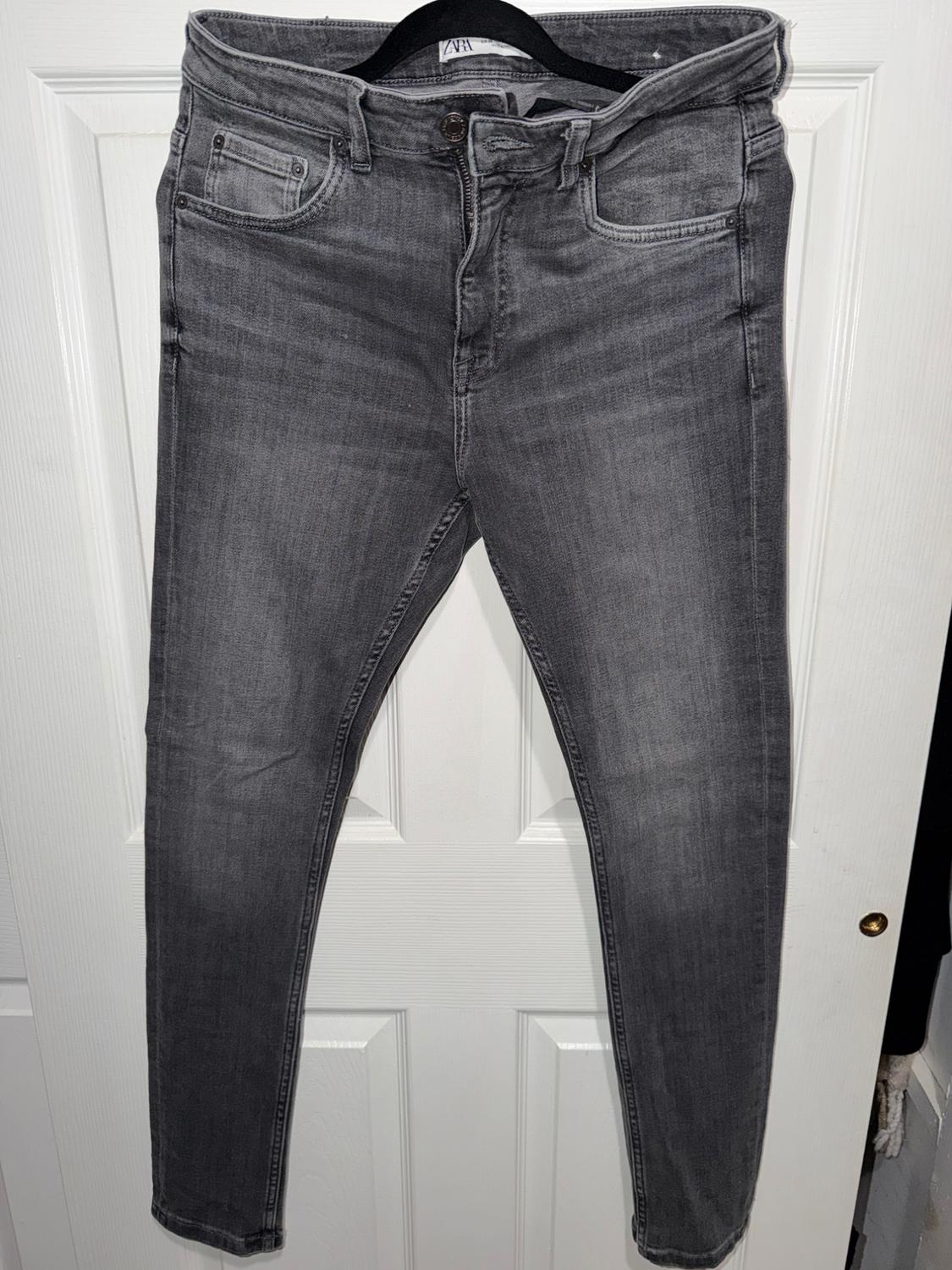 Grey jeans hanging on a white door, with a five-pocket design and button fastening. Visible label on the waistband.