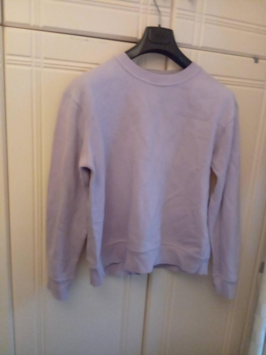 Lilac sweatshirt hanging on a black hanger in front of a beige wardrobe. The garment has a simple and casual design.