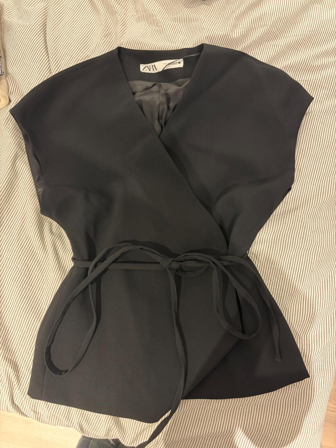 Black sleeveless blouse with belt, wrap style, on a striped fabric surface. Visible label with the text "ZARA".