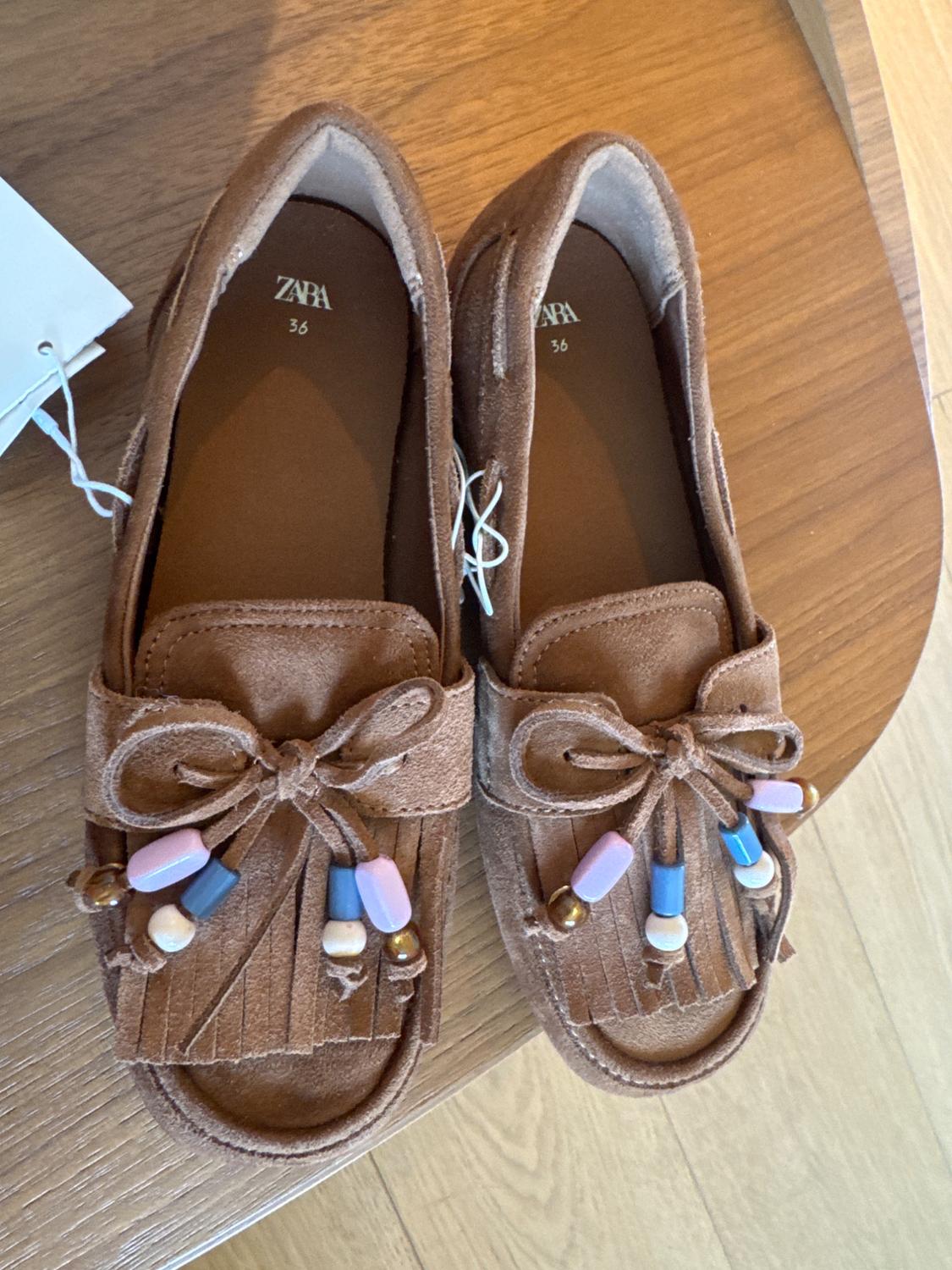 Brown loafers with tassels and colourful beads, "ZARA" brand and "36" size visible inside, on a wooden table.