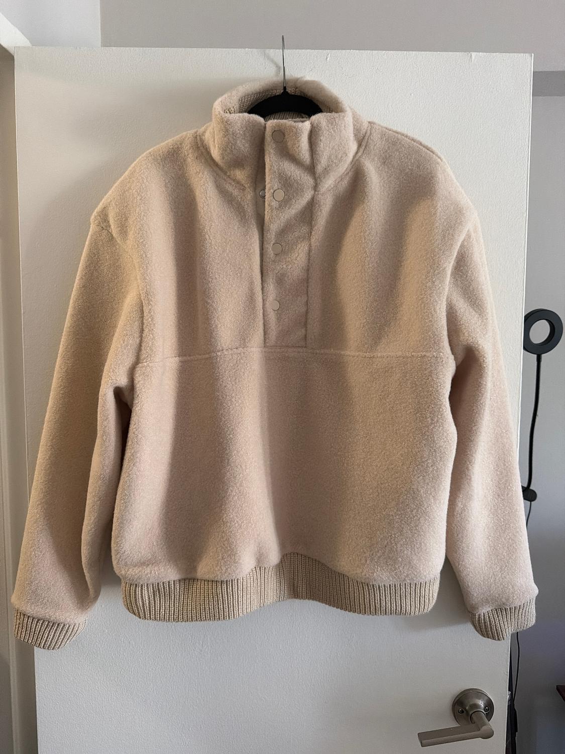 Beige wool roll-neck jumper with buttons, hanging on a hanger over a white door. Minimalist setting.
