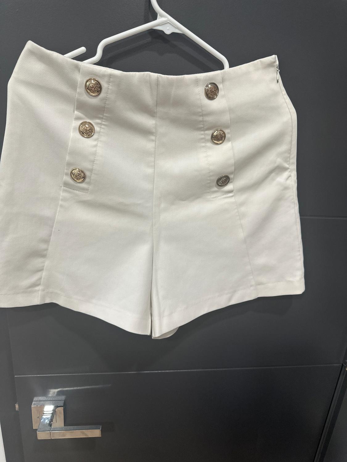 White shorts with six gold buttons on the front, hanging on a white hanger over a black door.