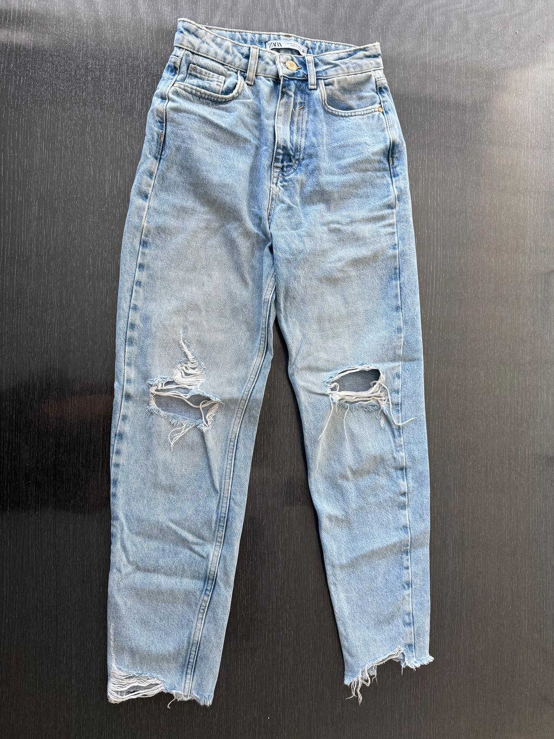 Distressed blue jeans with ripped knees and frayed hem, on a black background. Visible label at the waist.