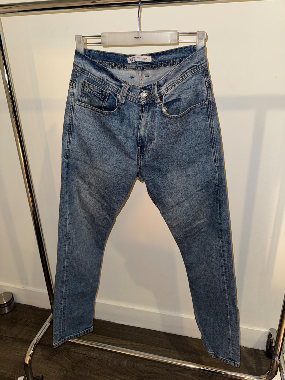 Light blue jeans hanging on a metal clothes rail with a white background. Visible label on the waist. Wooden floor.