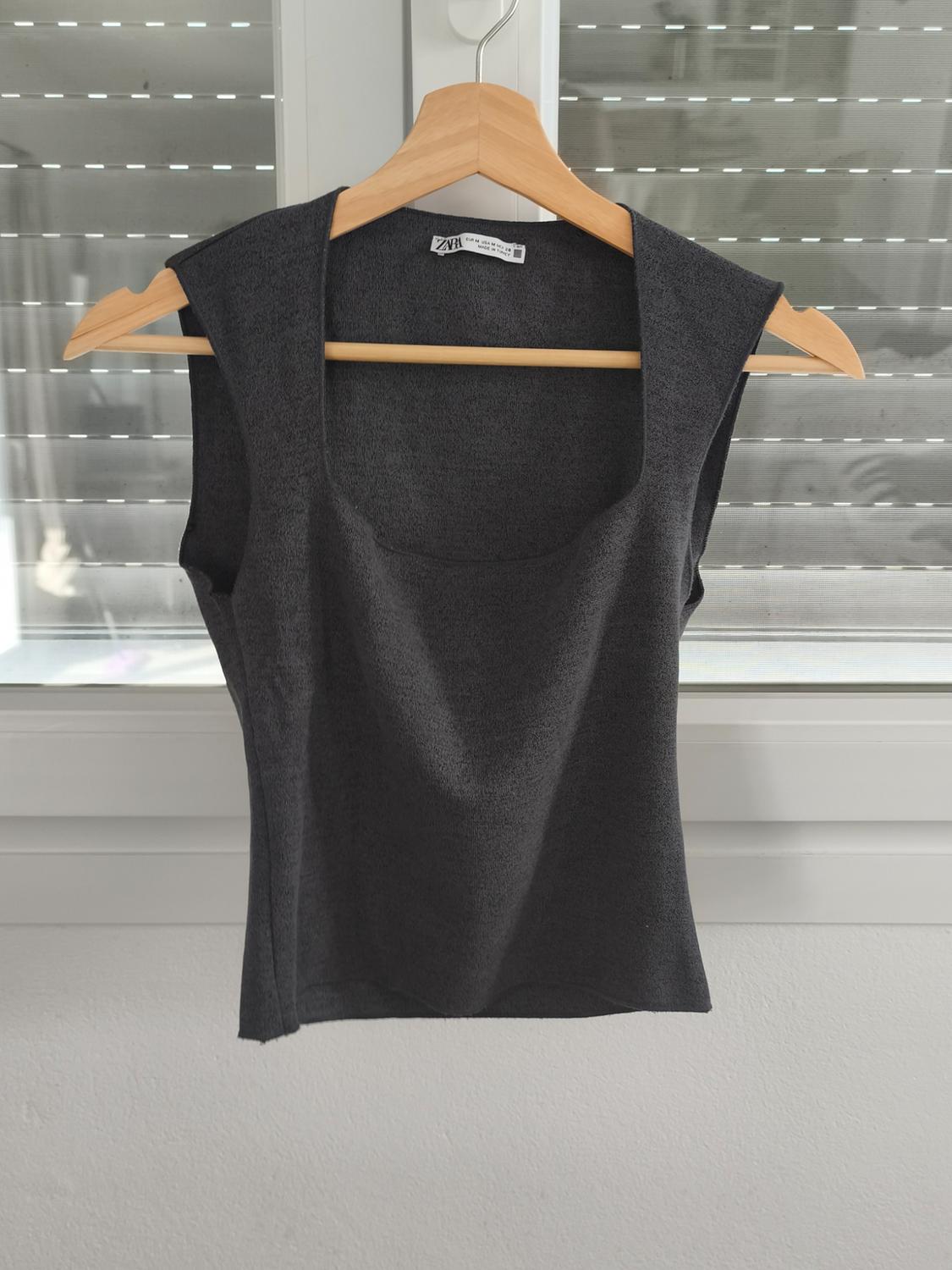 Dark grey sleeveless top hanging on a wooden hanger in front of a window with blinds. Label visible on the collar.