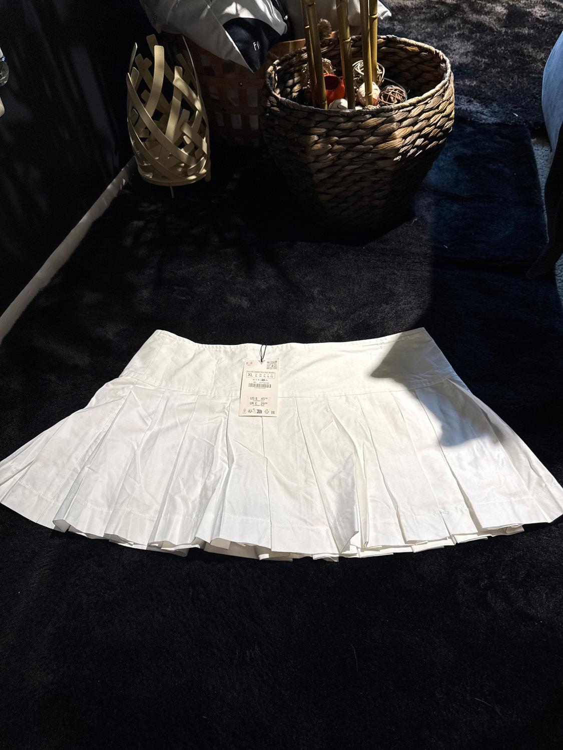 Pleated white skirt on a black rug, with a visible tag. In the background, decorative baskets and cushions in a dark setting.