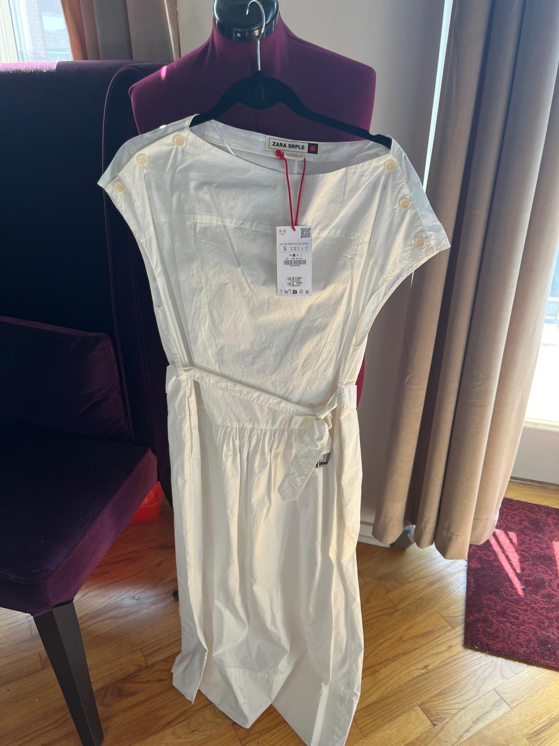 White dress hanging on a purple mannequin, with buttons on the shoulders and a visible tag. Background with curtains and wooden flooring.