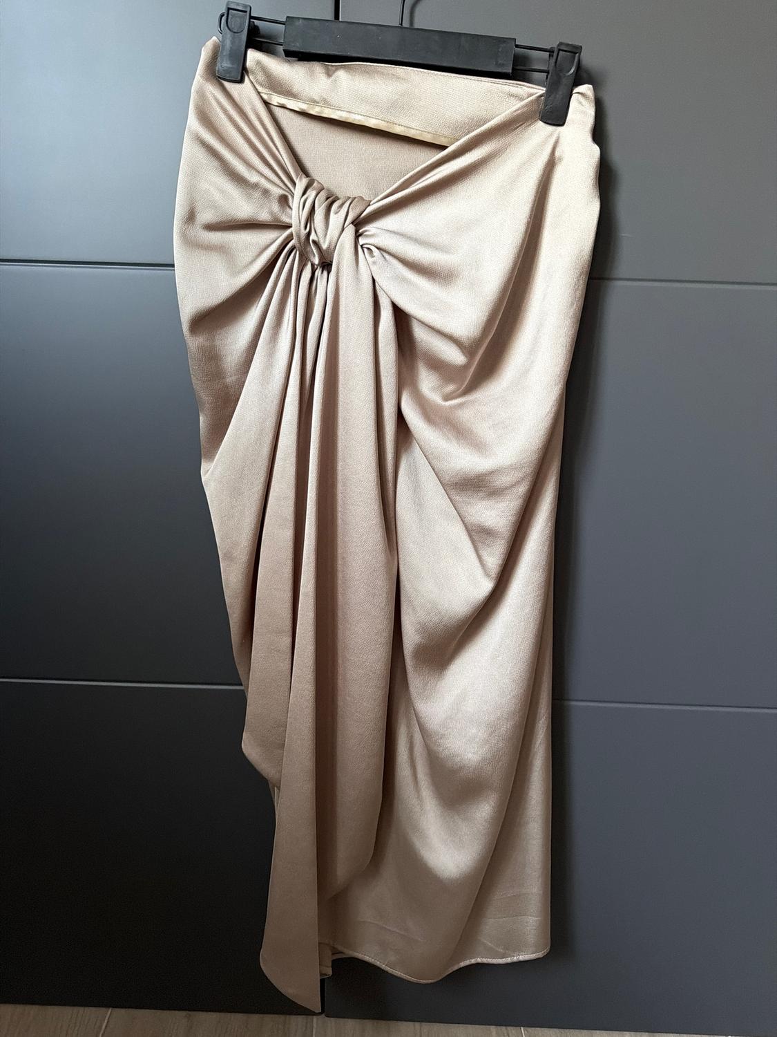 Beige draped skirt hanging on a black hanger against a dark grey background. The design features a decorative knot at the top.