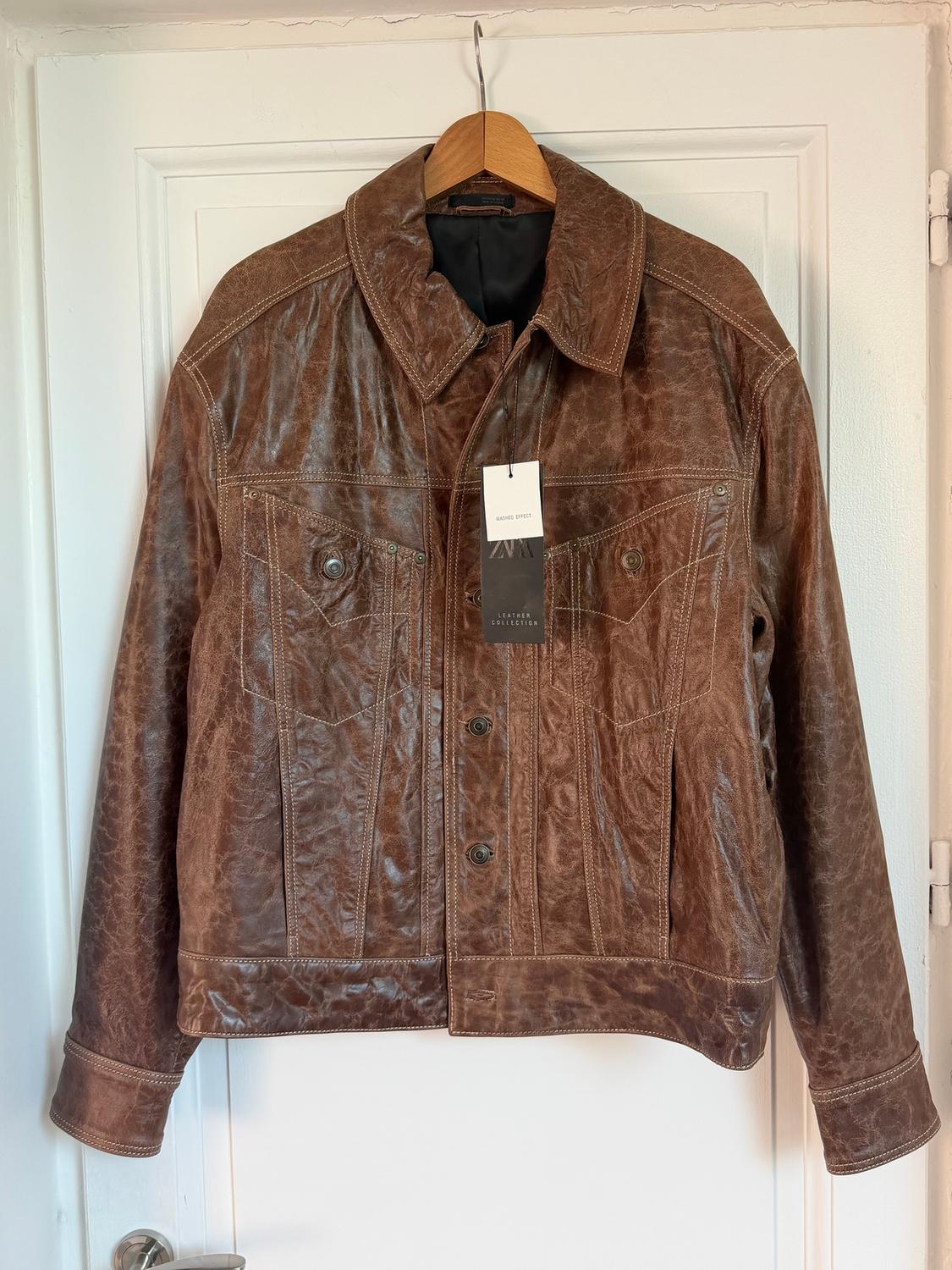 Brown leather jacket hanging on a wooden hanger, with a label that says "ZARA LEATHER COLLECTION" on a white door.