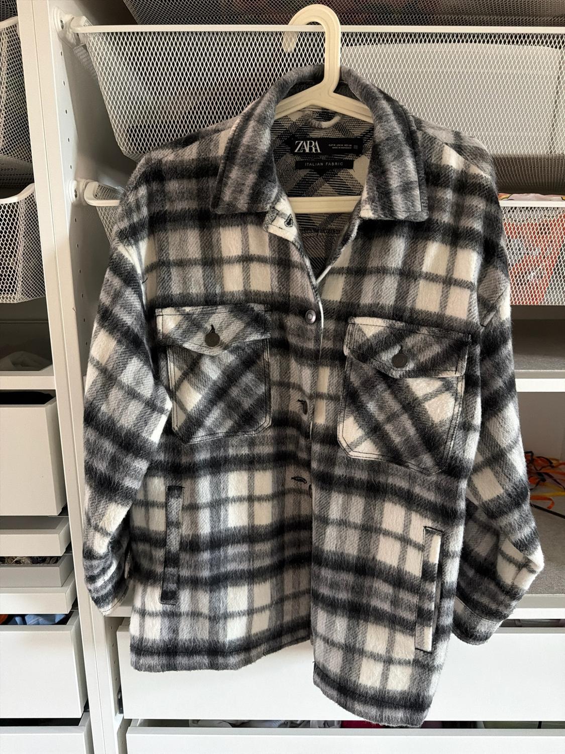 Black and white checked overshirt hanging in a wardrobe. Features front pockets and a label with the text ZARA ITALIAN FABRIC.