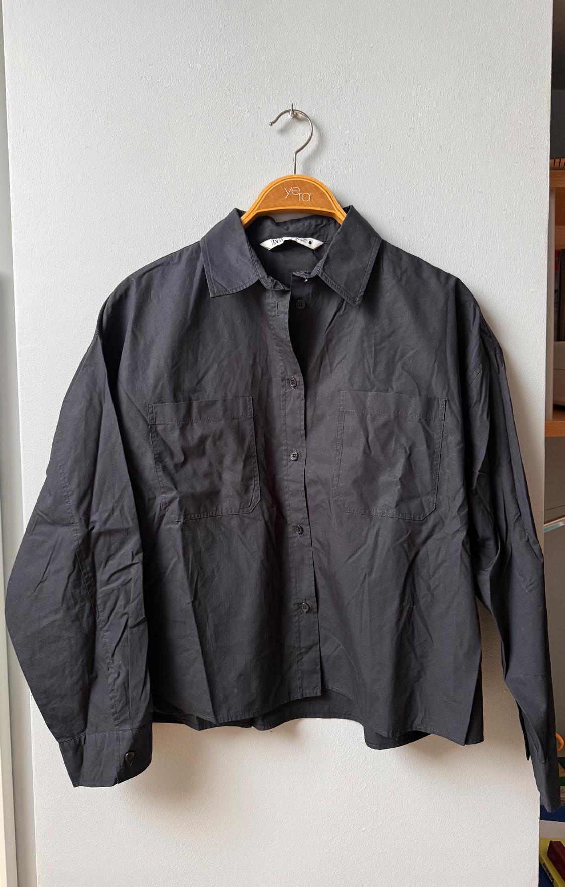 Black long-sleeved shirt with front pockets, hanging on a wooden hanger with the "Yera" brand visible.