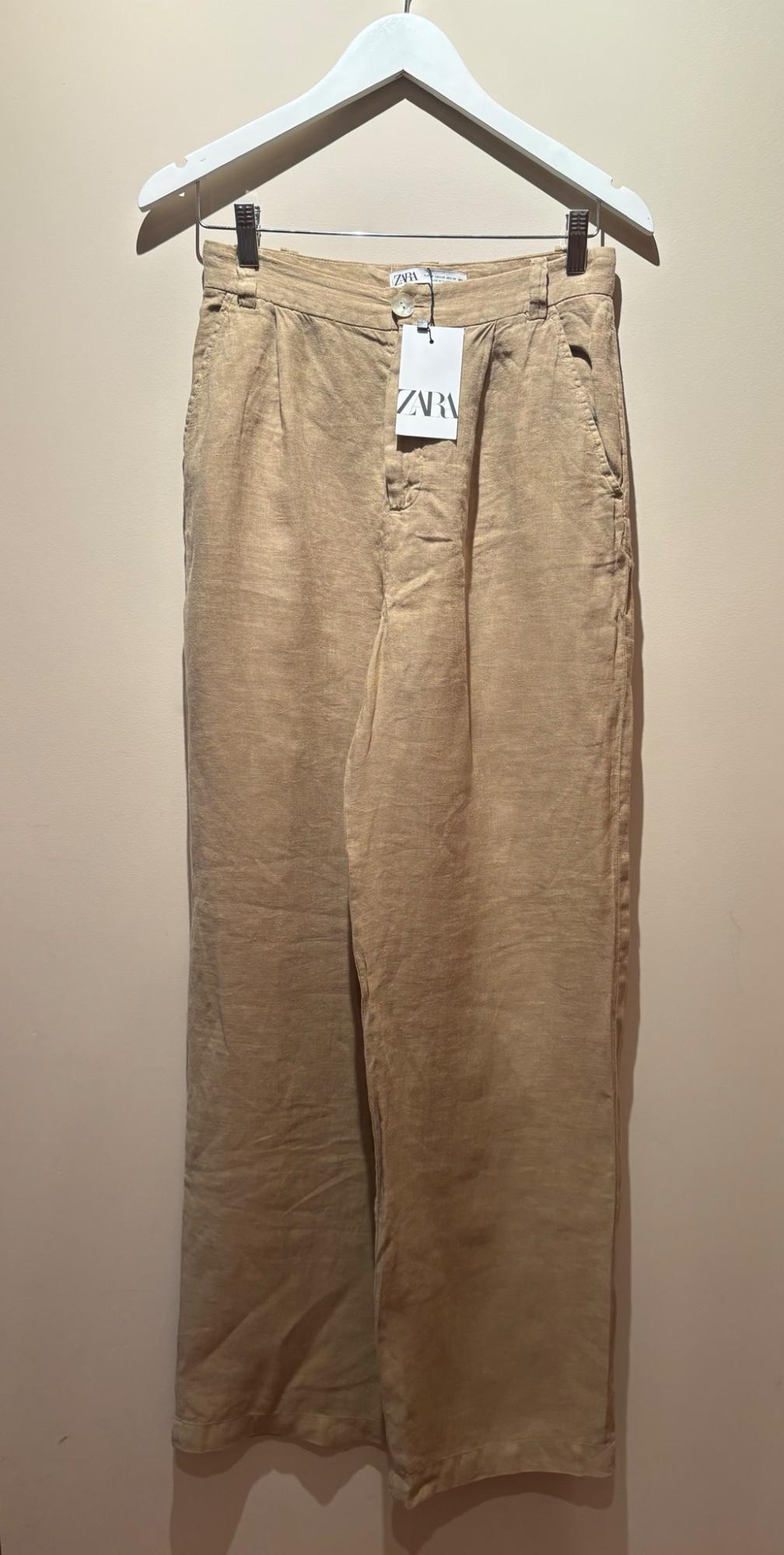 Beige trousers hanging on a white hanger against a light background. There is a label on the waistband with the text ZARA.