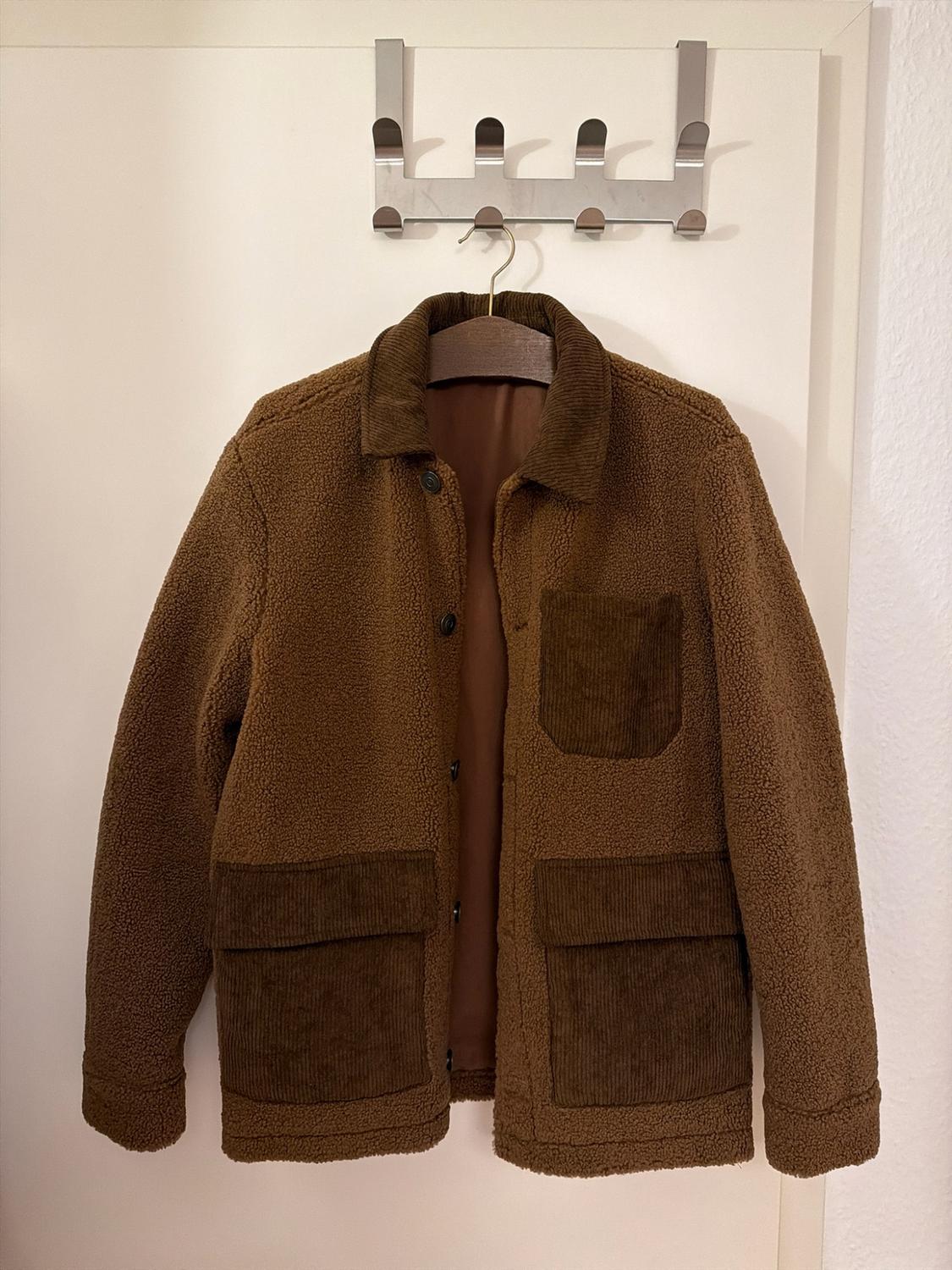 Brown jacket in curly fabric with a contrasting corduroy collar and pockets. Hanging from a hanger on a white door.