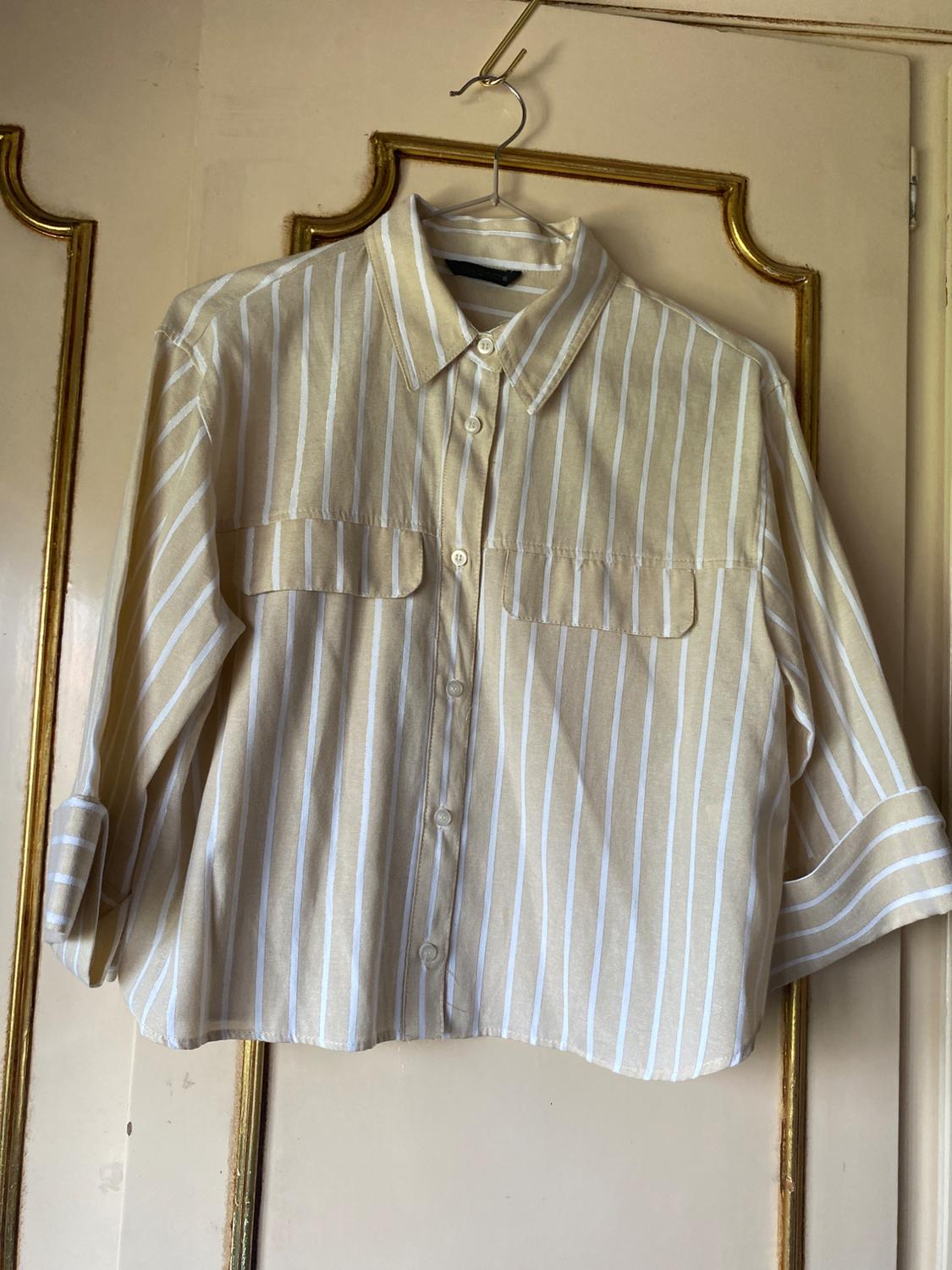 Beige and white striped shirt with front pockets, hanging on a hanger over a door with gold mouldings. Relaxed style.