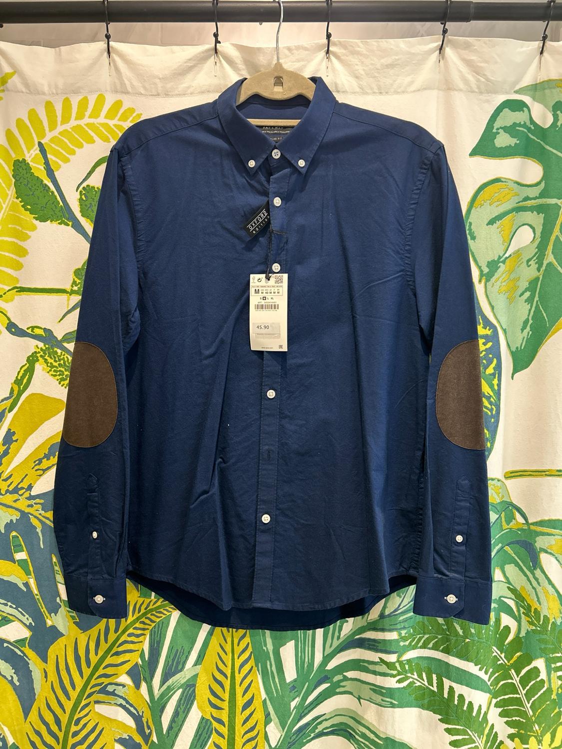Blue shirt with brown elbow patches hanging against a background of tropical leaves. Text: OXFORD edition, M, 45.90.
