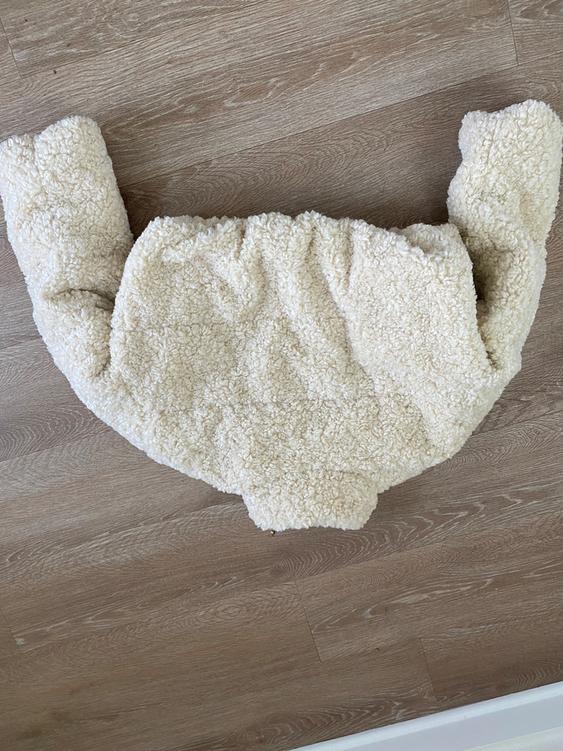 Beige plush jacket with a fluffy texture, placed on a light wooden floor. Sleeves extended to the sides.