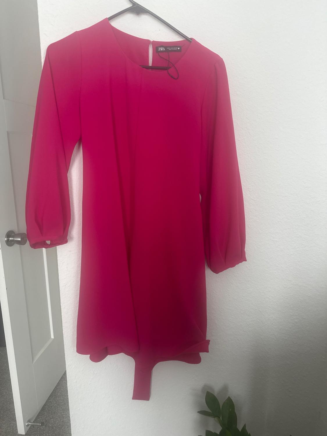Red dress with three-quarter sleeves hanging on a black hanger against a white wall. Asymmetric hem.