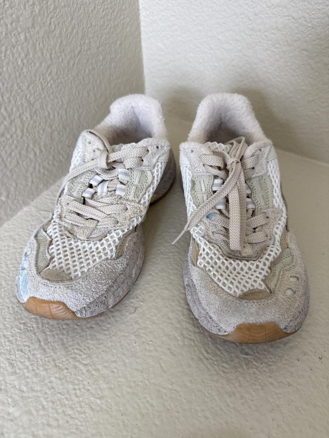 White sneakers with brown rubber soles and a mesh design, placed on a light surface next to a wall.
