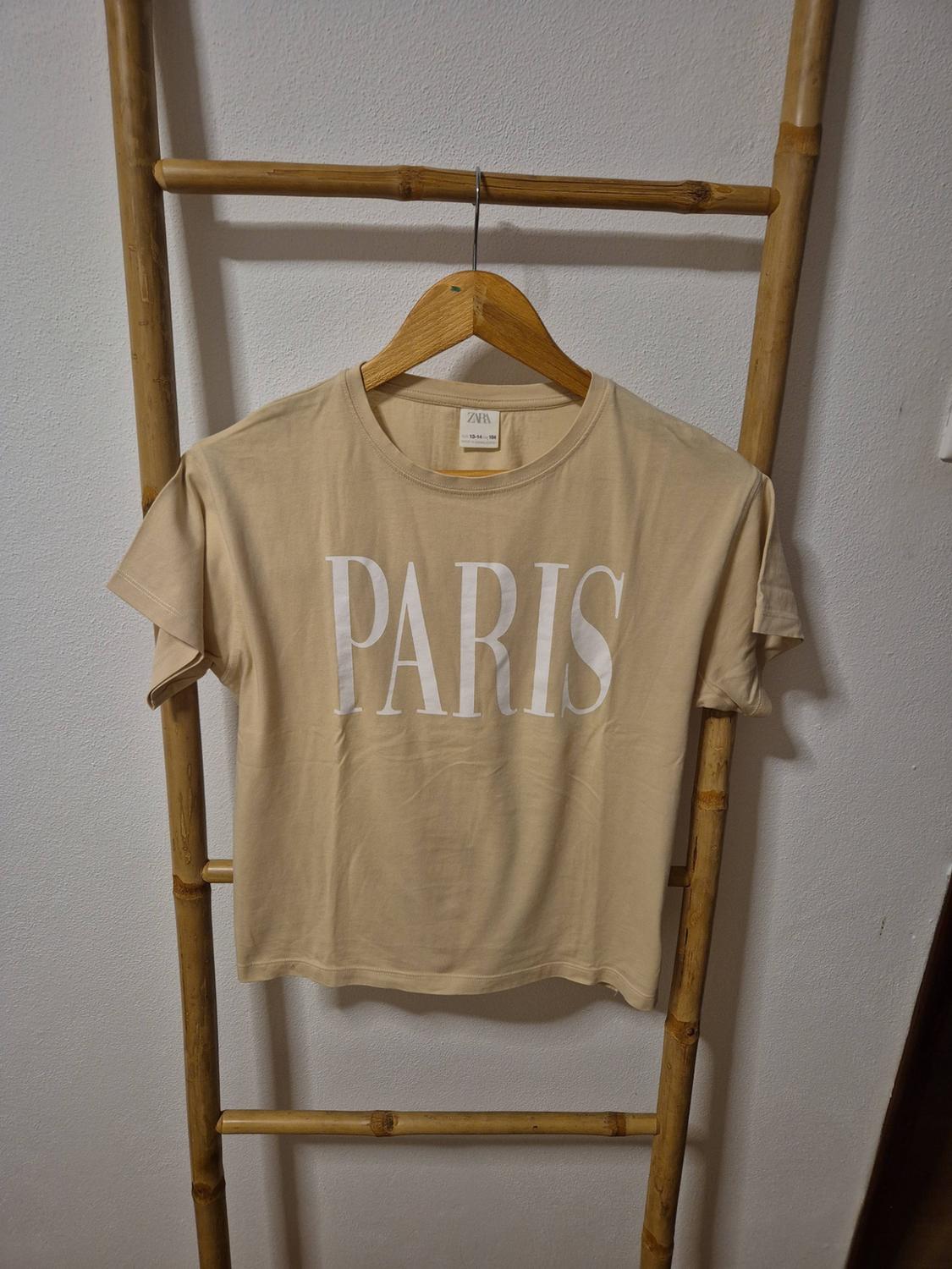 Beige short-sleeved T-shirt with the text PARIS in white, hanging on a wooden hanger over a bamboo ladder. ZARA label.