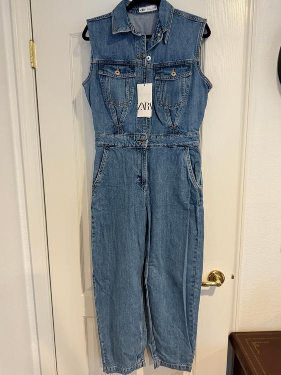 Sleeveless denim jumpsuit hanging on a white door. Visible tag with the text "ZARA". Front pockets and button closure.