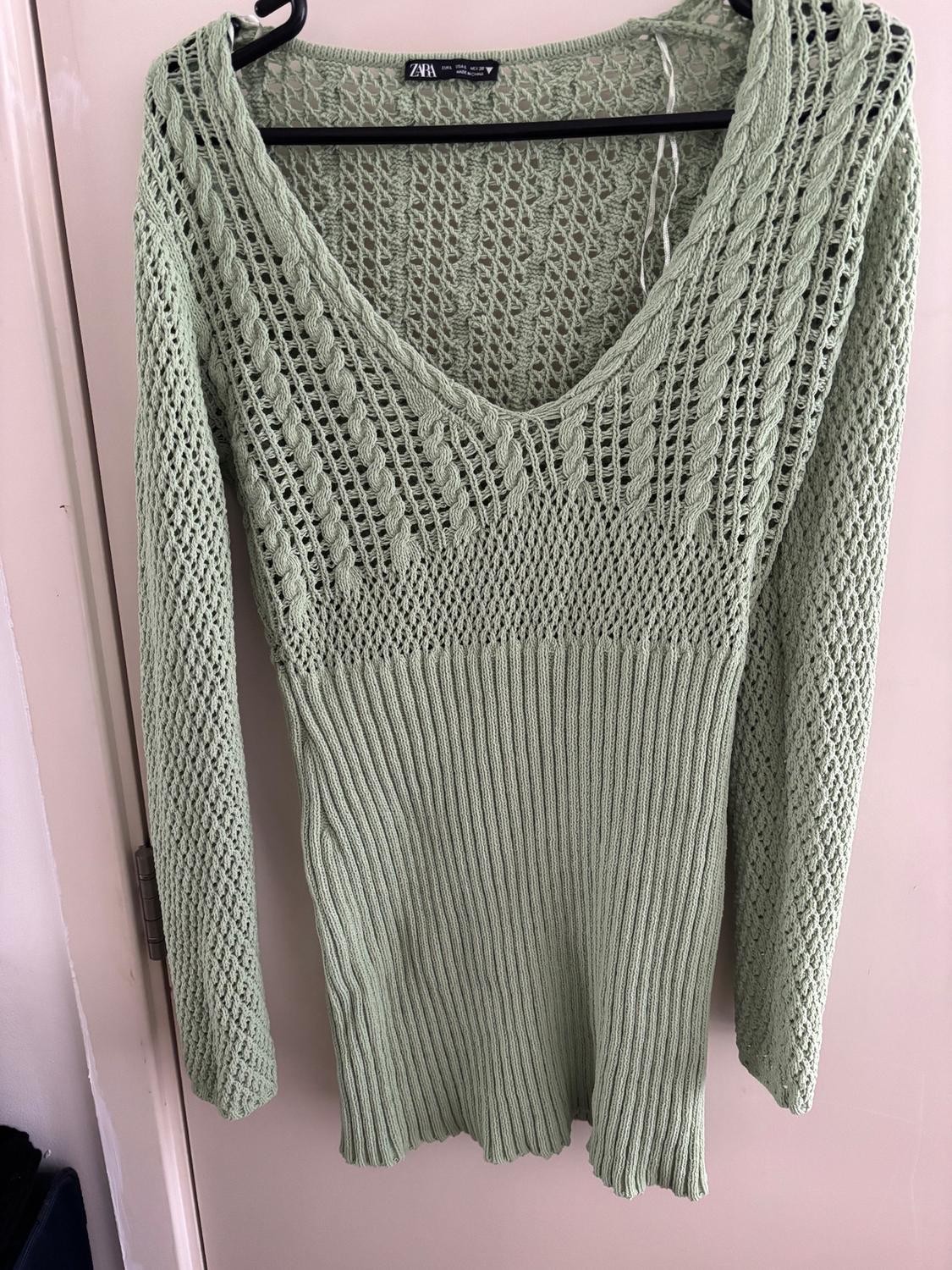 Light green open-knit jumper with a V-neck and long sleeves, hanging on a black hanger over a white door.