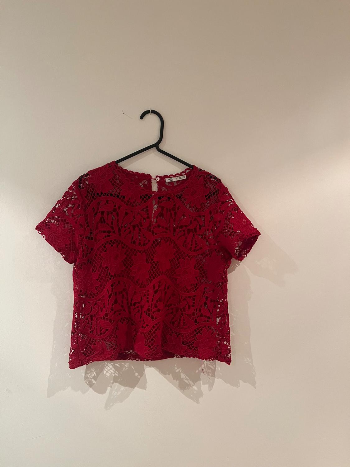 Red lace blouse with a floral design hanging on a black hanger against a white wall.