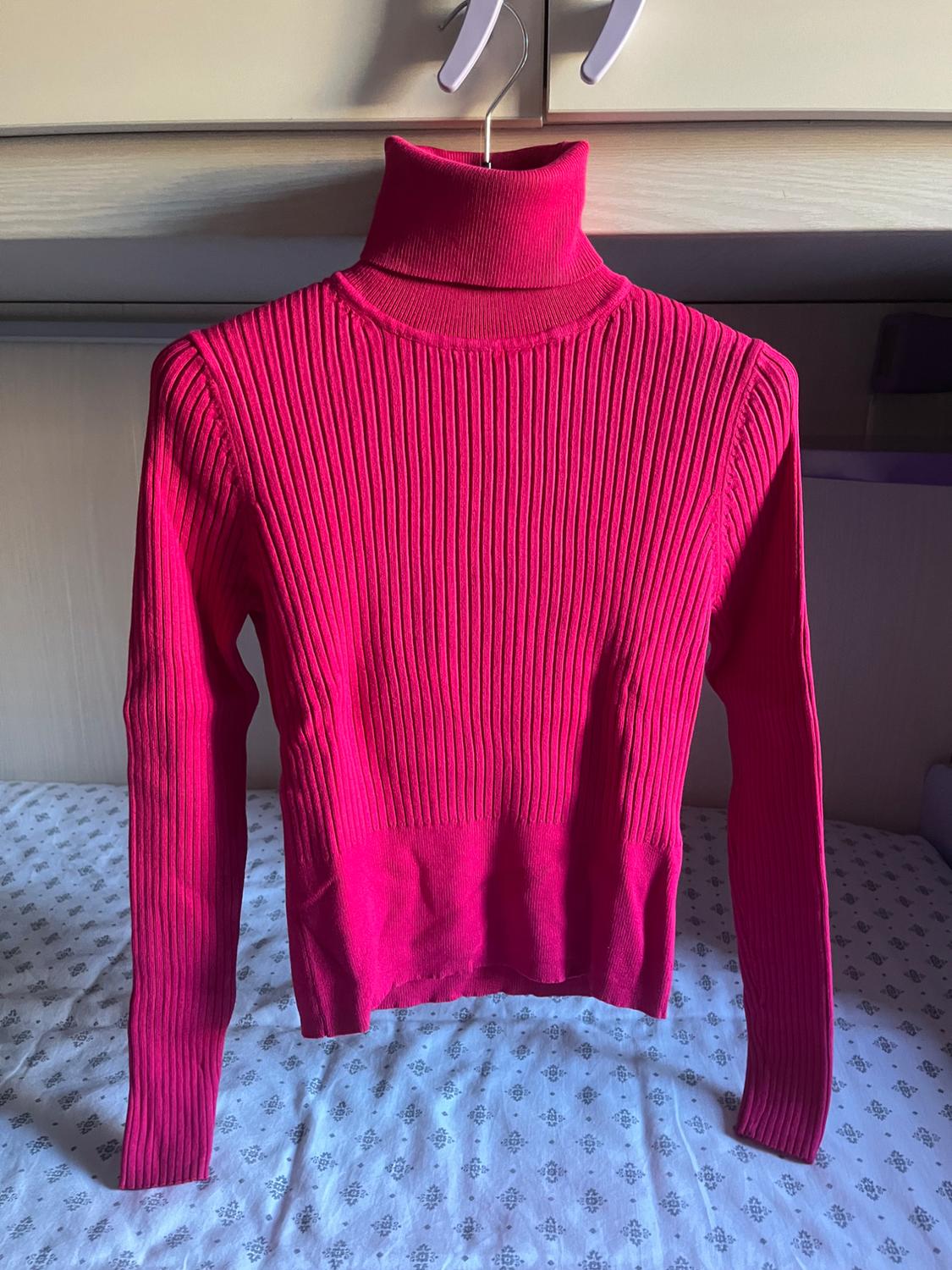 Fuchsia pink ribbed knit roll-neck jumper hanging on a hanger over a bed with patterned sheets. Relaxed and casual atmosphere.