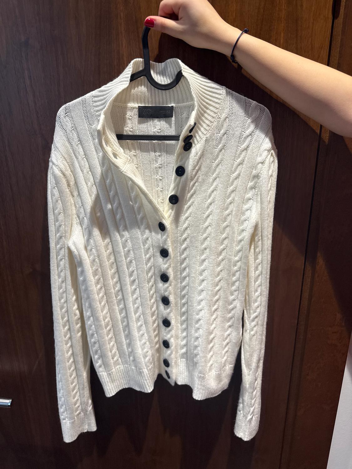White knitted cardigan with black buttons, hanging on a hanger. Hand with red painted nails holding the hanger.