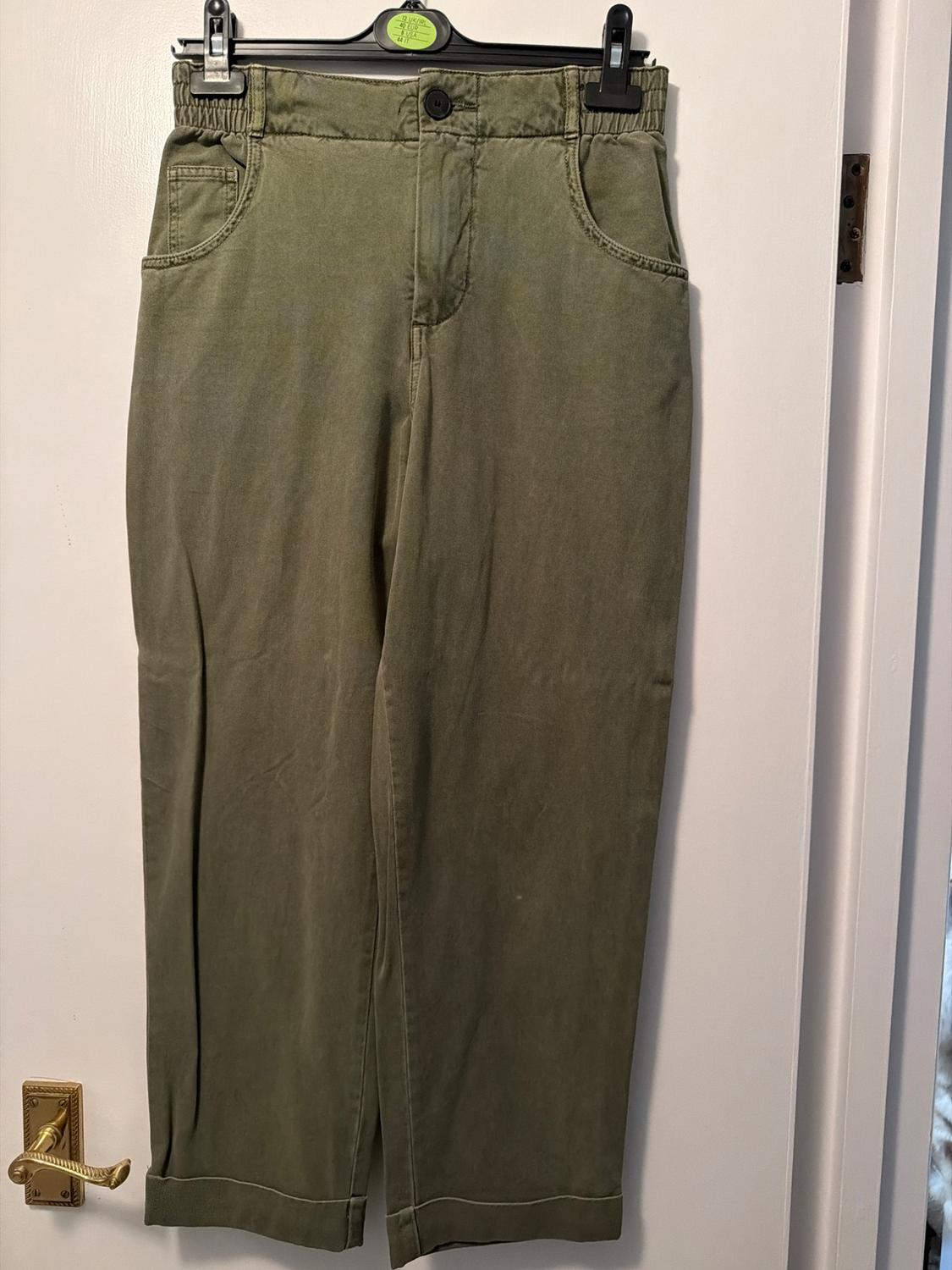 Green trousers hanging on a black hanger over a white door. They have an elasticated waist and side pockets.