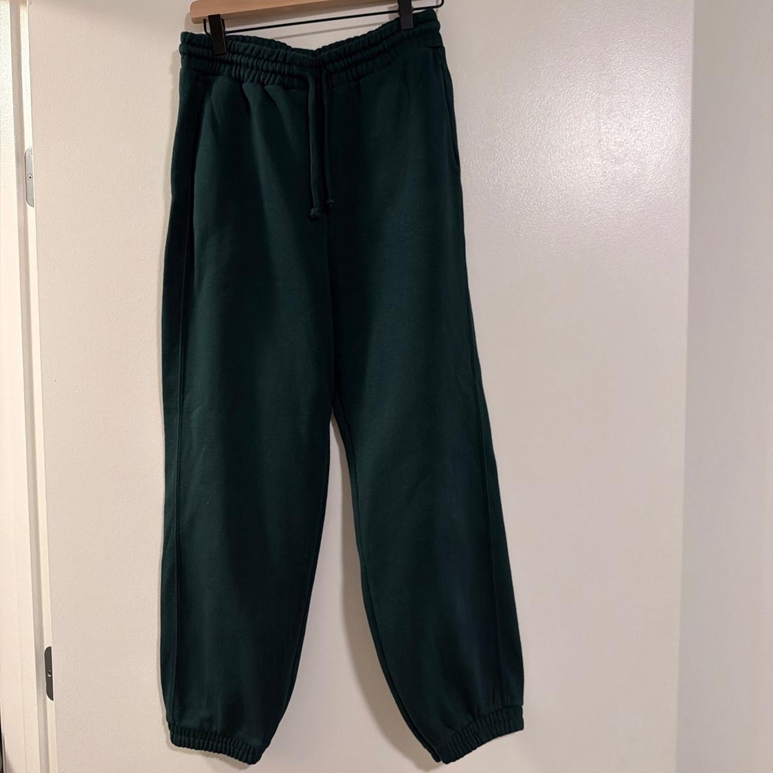 Dark green jogging bottoms hanging on a wooden hanger against a white wall. Featuring an elasticated drawstring waist and cuffed hems.