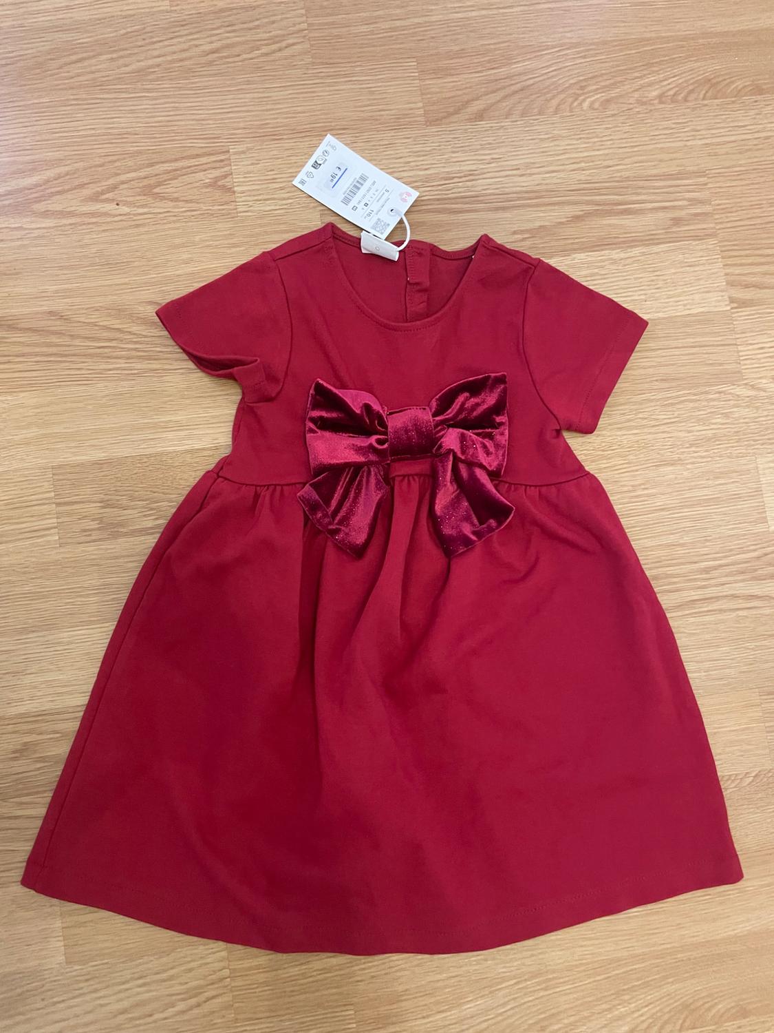 Short-sleeved red dress with a large velvet bow in the centre, laid out on a wooden floor. Label on the neck.