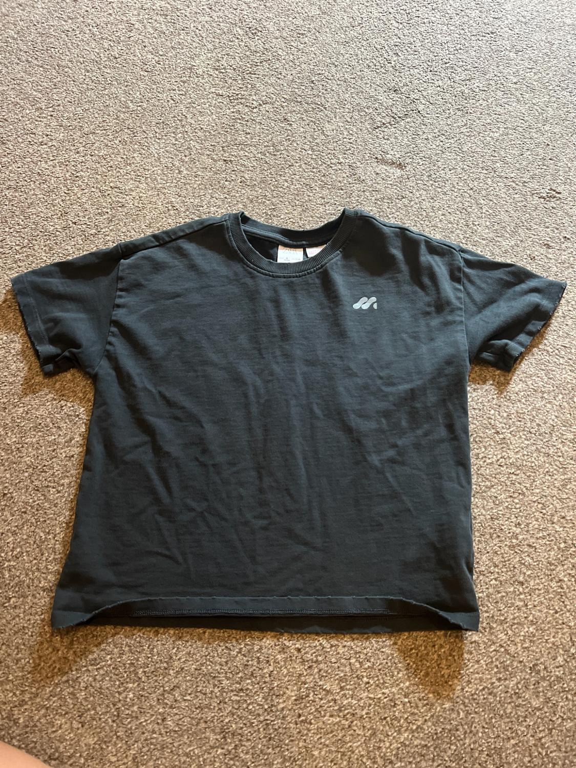 Dark grey short-sleeved T-shirt with a small white logo on the chest, on a textured brown rug.