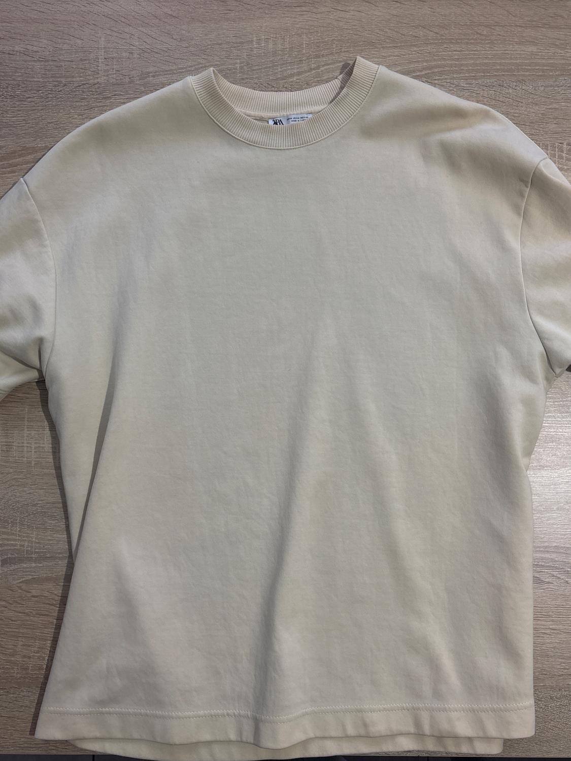 Beige short-sleeved, crew-neck t-shirt on a light wood background. Visible neck label.