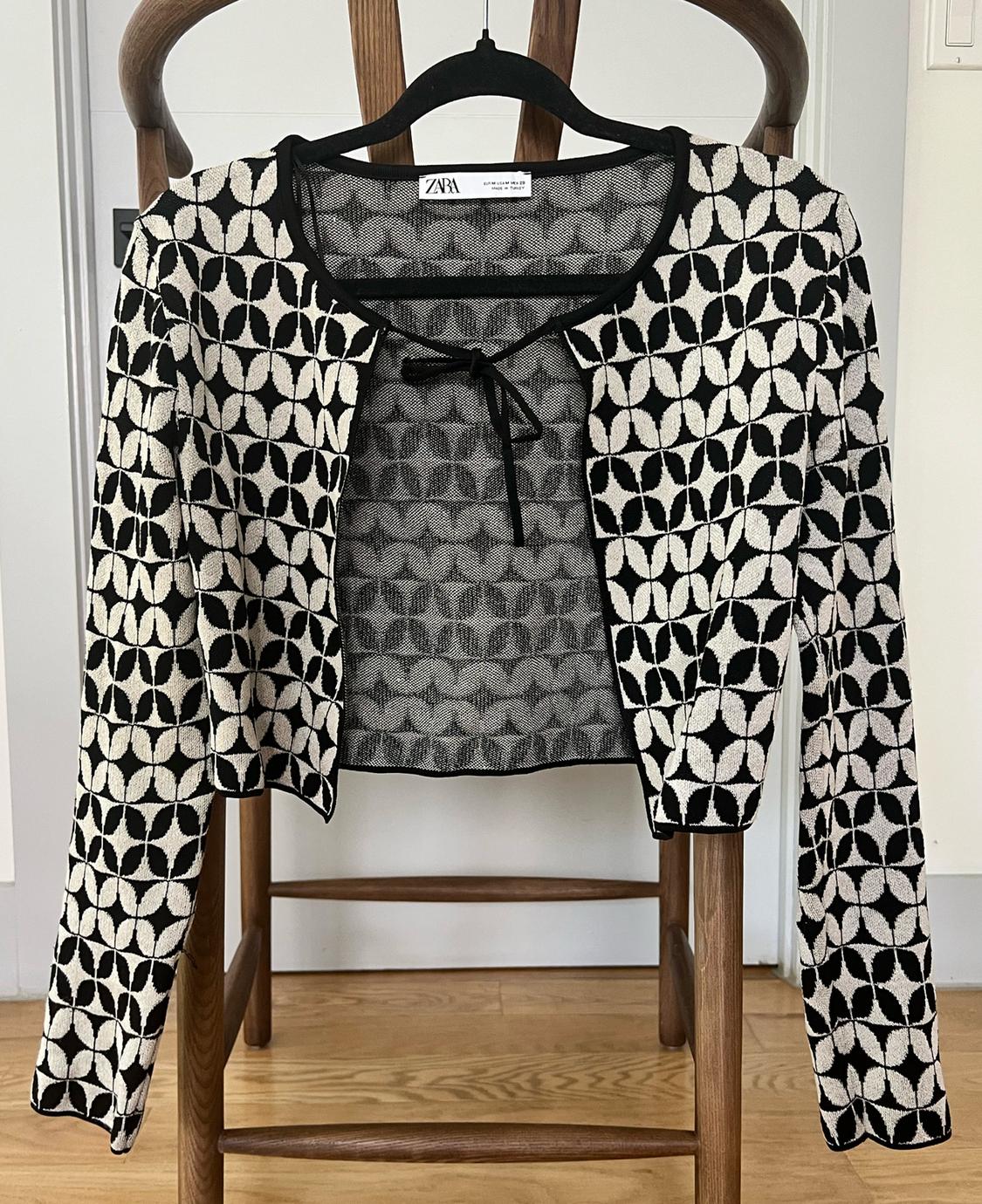 Short black and white patterned jacket with geometric design, neck tie, hanging on a hanger on a wooden chair.