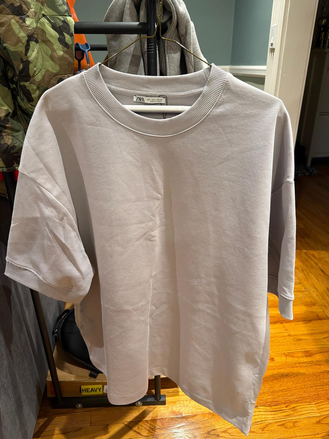 Light gray crew-neck T-shirt with short sleeves hanging on a hanger, with a Zara label. Below, a box with the word HEAVY visible.