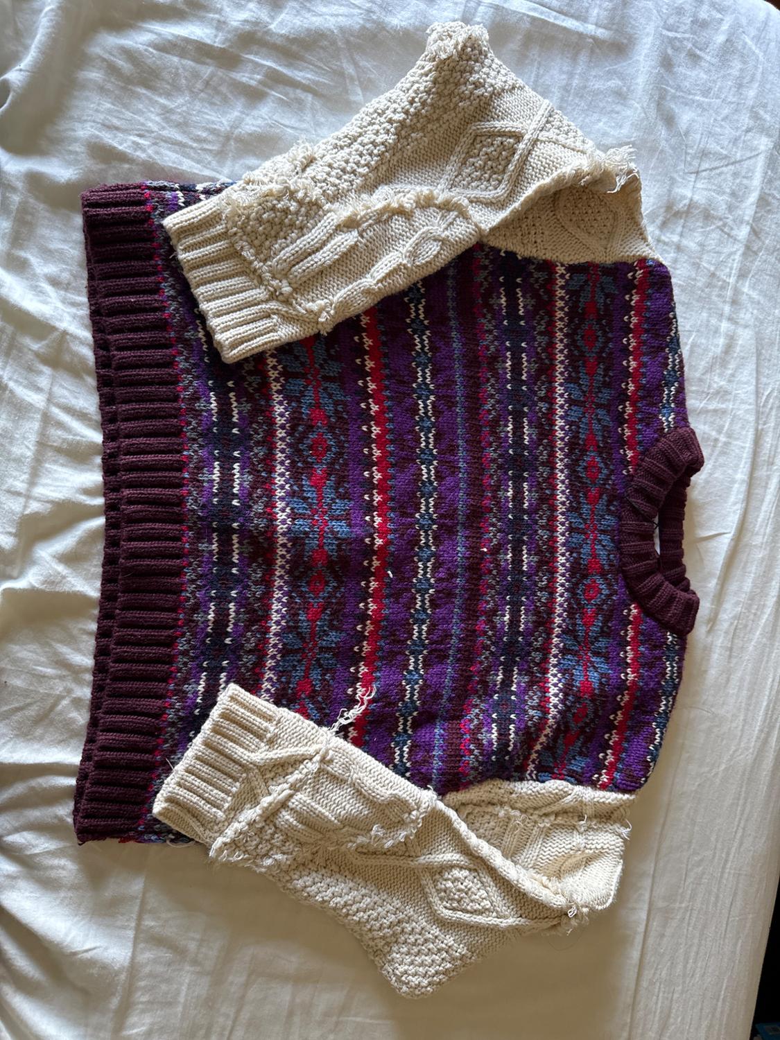 Knit sweater with a body of geometric patterns in shades of purple and red, and cable knit sleeves in cream color, on a white bed.