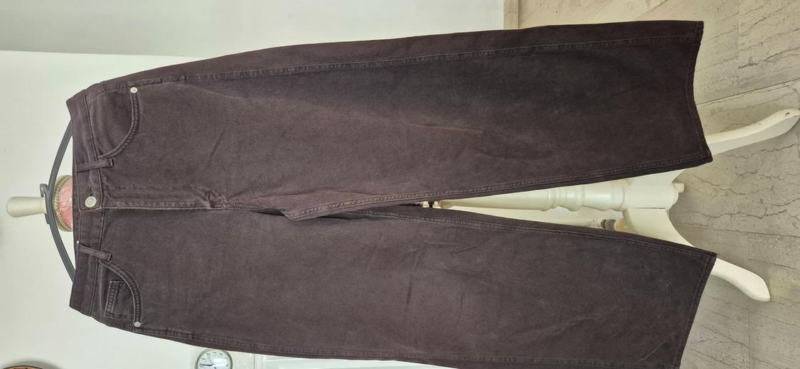Dark brown baggy jeans hanging on a hanger in front of a white wall and a white pedestal.