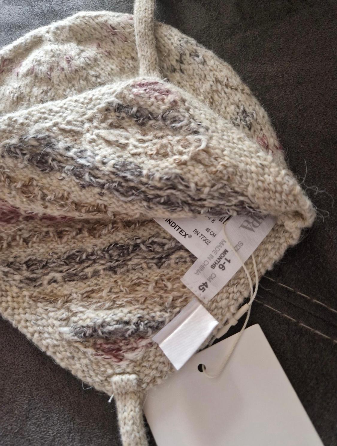 Knitted woolly hat in beige, grey, and pink tones on a dark background. Visible labels with text: "INDITEX", "MADE IN CHINA", "SIZE 1-6 MONTHS".