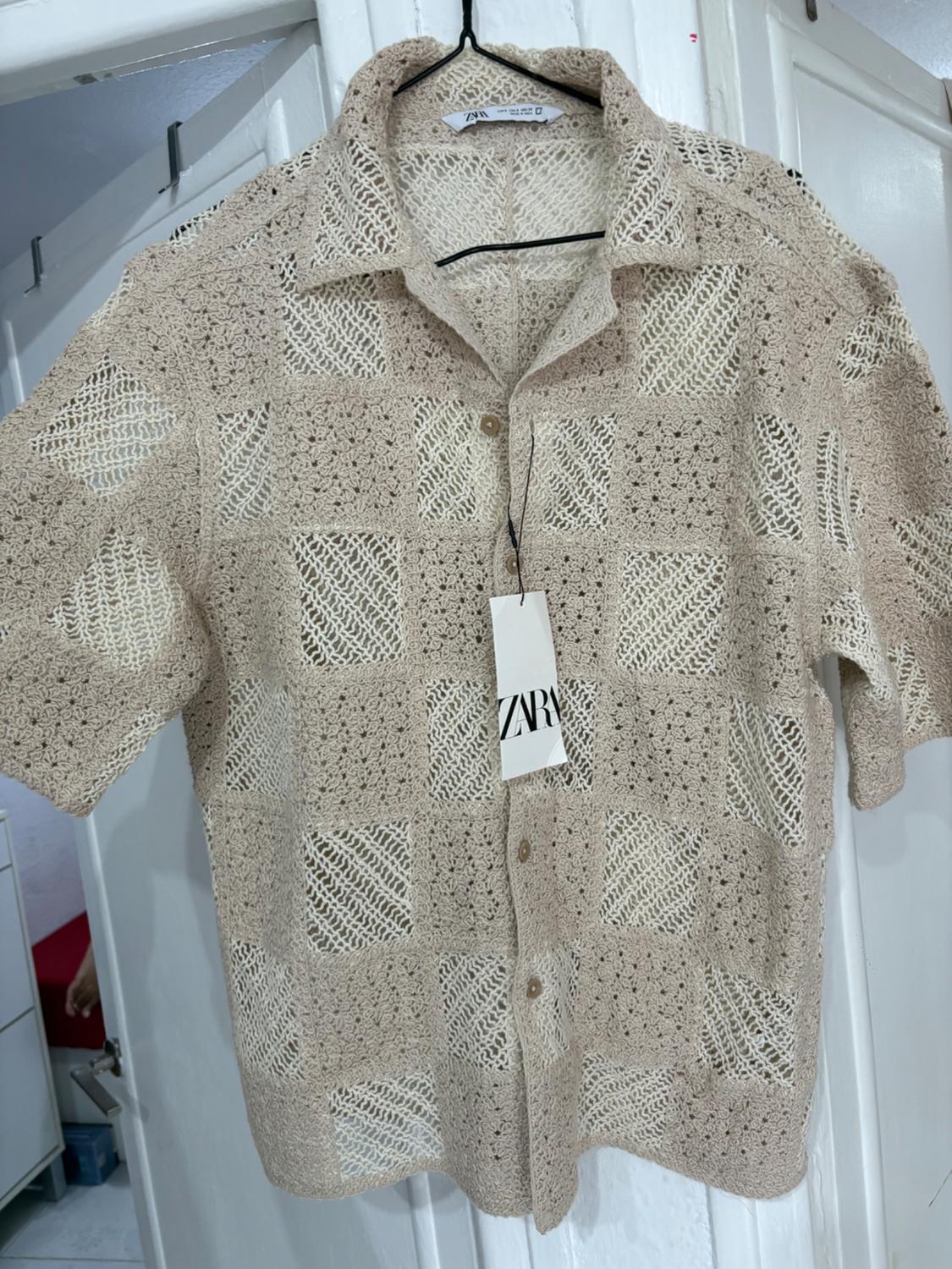 Beige crochet shirt with a check pattern hanging on a hanger. Label visible with the text "ZARA". Background of white doors.