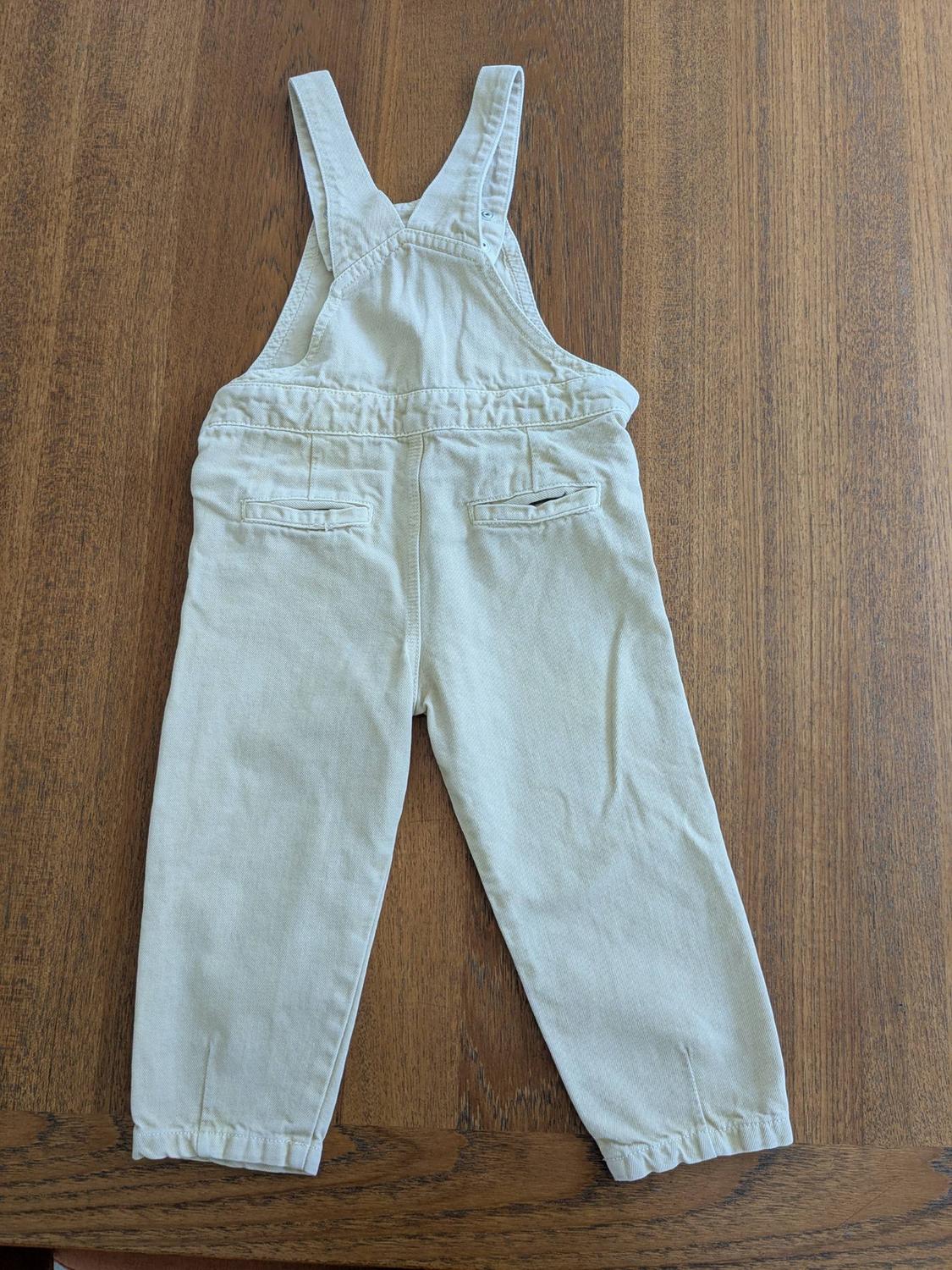 Light-colored fabric overalls in beige with straps, back pockets, and a straight cut, on a wooden table.