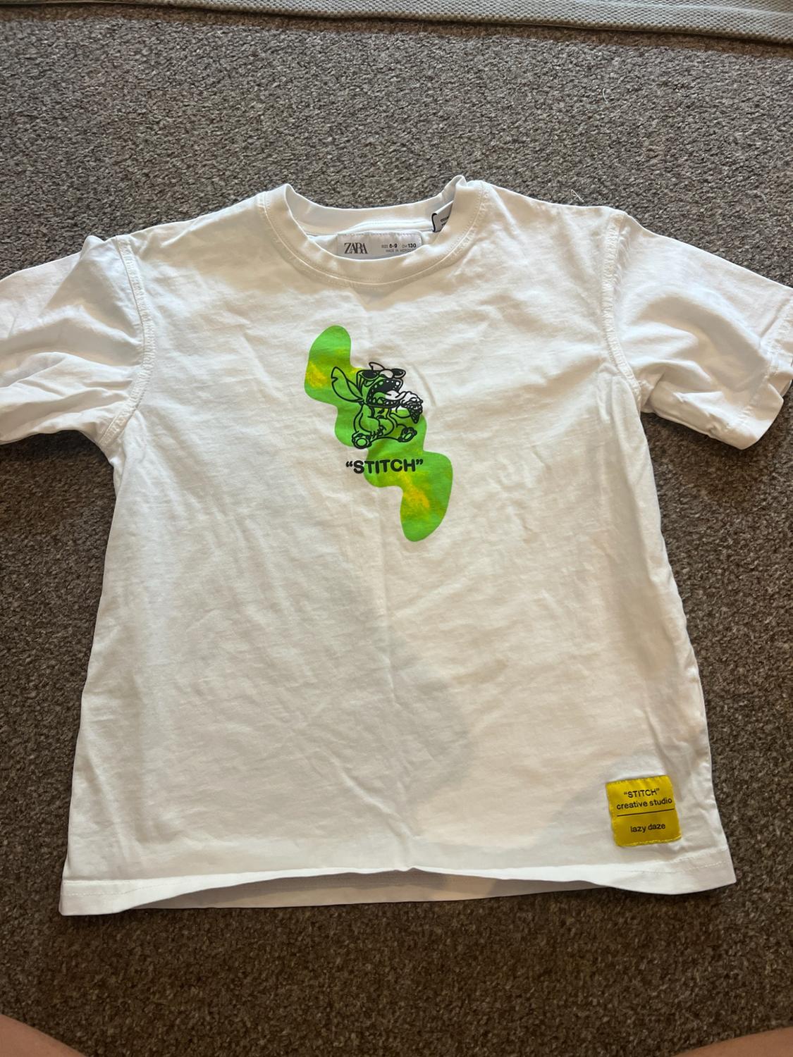 Short-sleeved white t-shirt with a green graphic and the text "STITCH". Yellow patch with the text "STITCH" creative studio lazy daze.