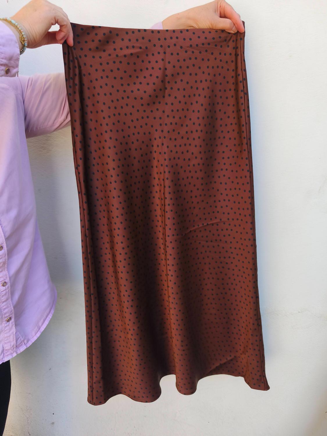Person holding a brown skirt with black polka dots. Wearing a lilac shirt and gold bracelet. White background.