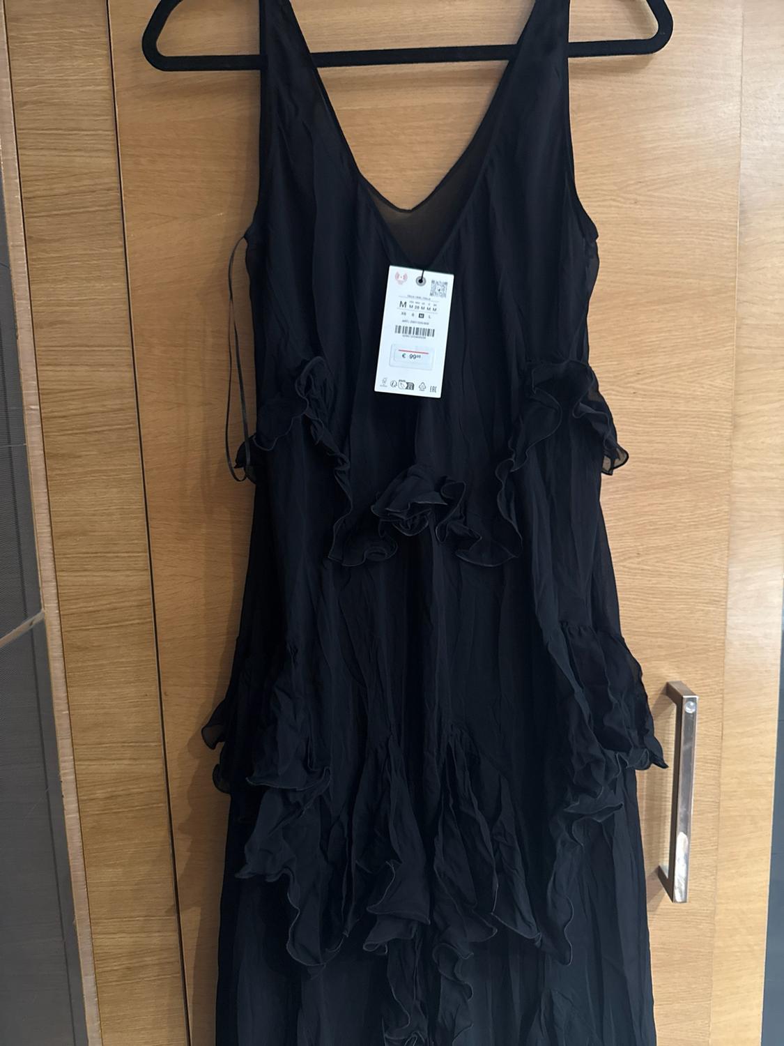 Black dress with frills and a V-neck, hanging on a wooden door. Visible label with the text M and €99.