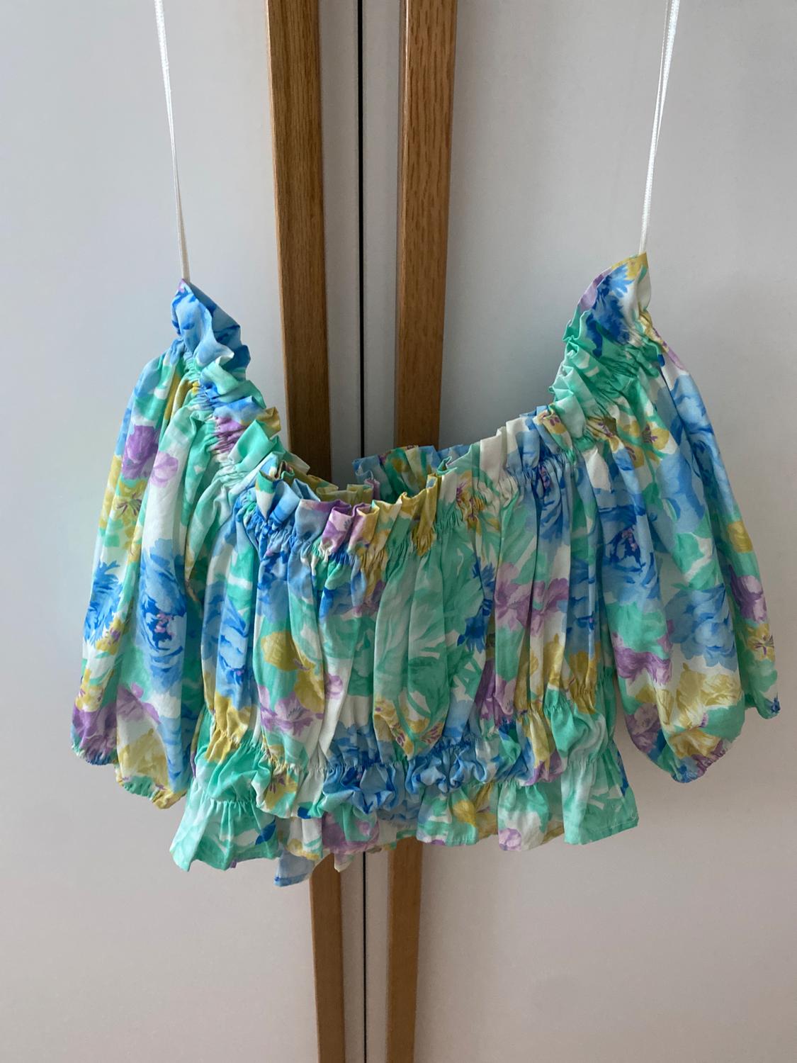 Cropped top with a pastel floral print hanging on a white hanger against a white and wood wardrobe background.