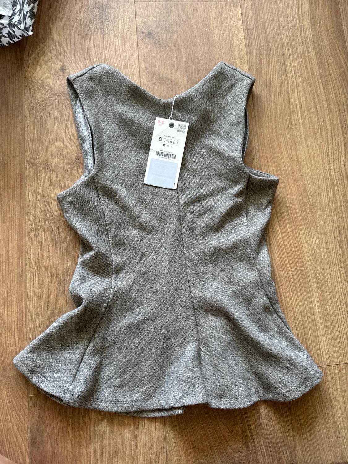 Grey sleeveless knitted peplum hem top on a wooden floor. Tag with text S S 26 S S P.