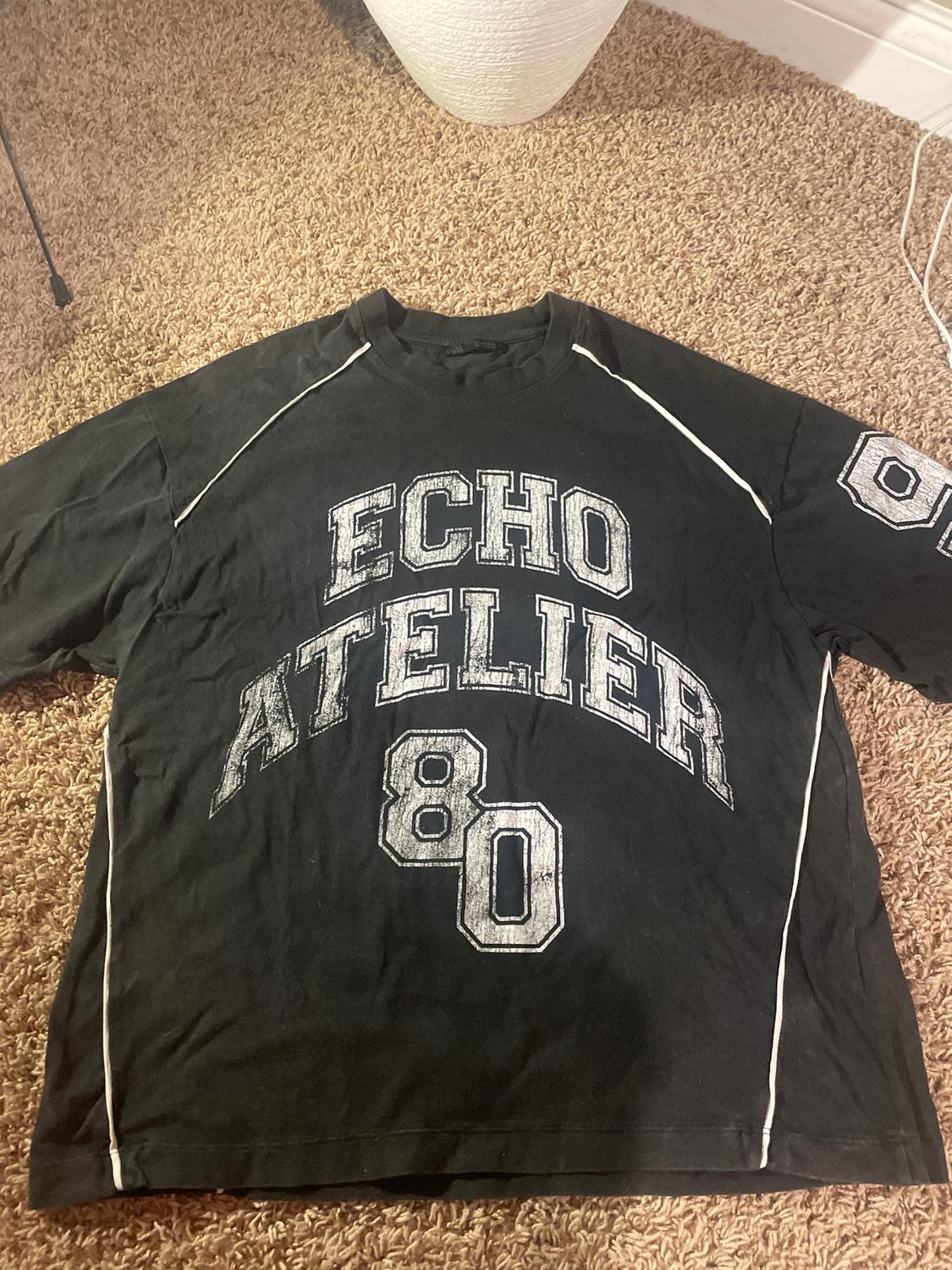 Black T-shirt with "ECHO ATELIER 80" text in distressed white letters, on a brown rug.